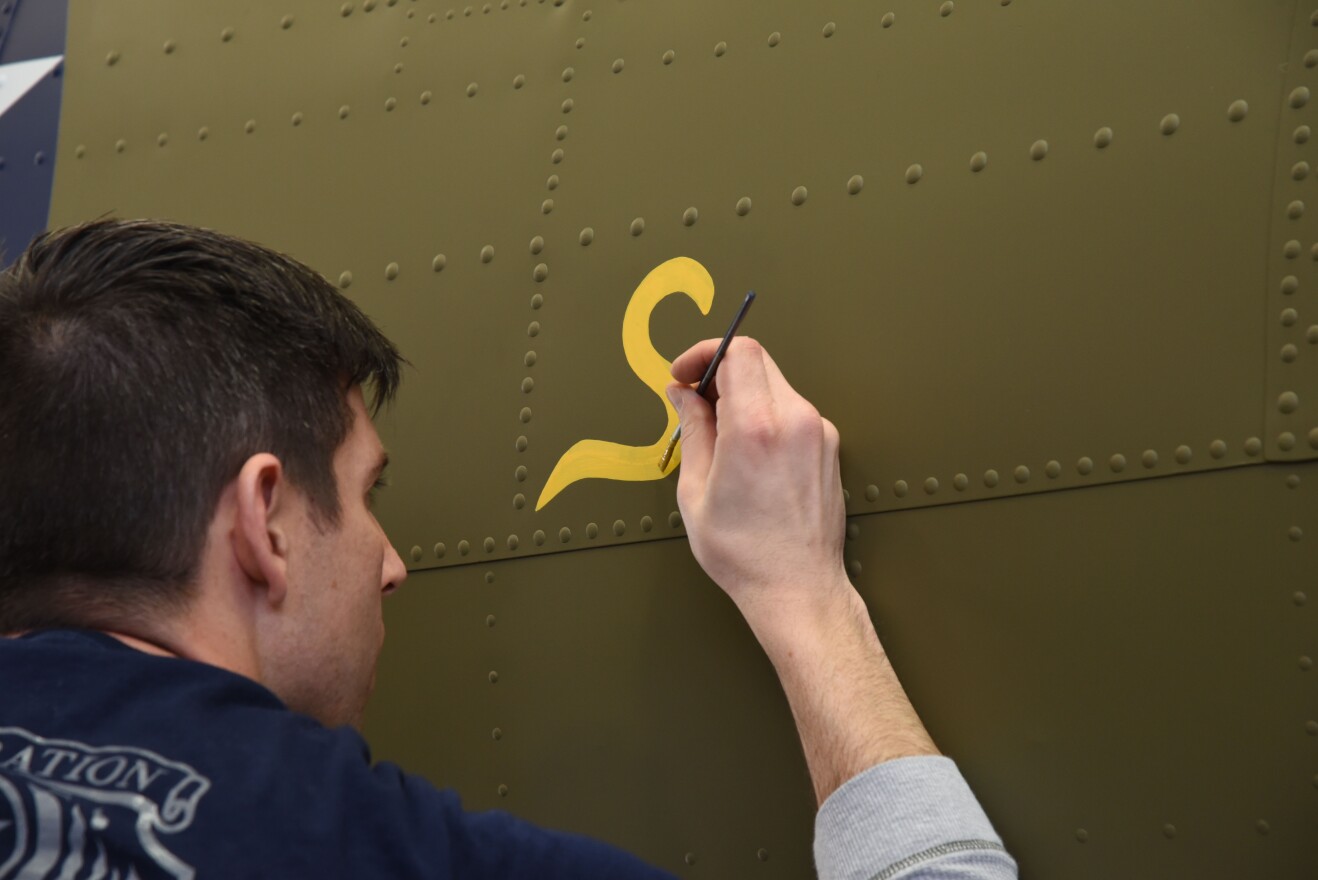 Museum restoration specialist Casey Simmons paints the letter "S" for "Sally" below the left waist gun position of the Boeing B-17F Memphis Belle as part of the aircraft's restoration.